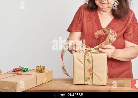 Donna matura ispanica che fa un arco con un nastro decorativo per decorare un regalo di Natale. Design luminoso con spazio copia. Concetto di holida di imballaggio Foto Stock