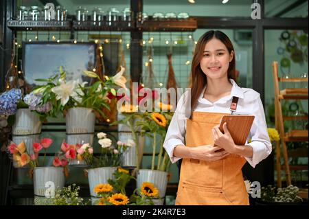 Ritratto, bella e affascinante giovane fiorista asiatico o imprenditore fioraio in un grembiule si trova di fronte al suo ingresso negozio di fiori, holdi Foto Stock