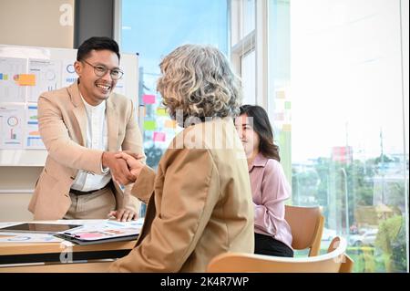 Uomo d'affari asiatico professionale e allegro che si stringe la mano con dirigente senior femminile nella riunione. Saluto, Congratulazioni Foto Stock