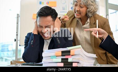 Stressato e frustrato imprenditore asiatico millenario o dipendente maschio ha ricevuto reclami dal suo capo. Arrabbiato e pazzo capo femmina senior sta urlando un Foto Stock