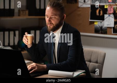 Investigatore che lavora in ufficio utilizzando un computer, processo di analisi delle prove di un caso penale, investigatore beve caffè di notte Foto Stock