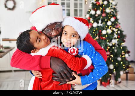 Papà e bambini - una figlia e un figlio vestiti in costume di Babbo Natale con una falsa barba che abbraccia e sorride sullo sfondo dell'albero di Natale. Foto Stock