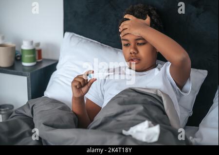 Ragazzo malato sdraiato a letto con febbre e controllo della temperatura Foto Stock