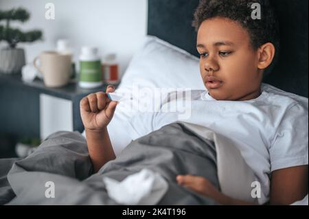 Ragazzo malato sdraiato a letto con febbre e controllo della temperatura Foto Stock