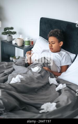 Ragazzo malato sdraiato a letto con febbre e controllo della temperatura Foto Stock