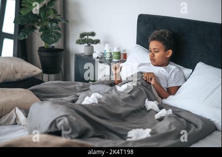 Ragazzo malato sdraiato a letto con febbre e controllo della temperatura Foto Stock