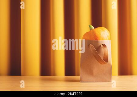 Sfondo decorazione autunnale con piccola zucca gialla in borsa shopping kraft su tavolo di legno contro tende gialle. Vista frontale, spazio di copia. Foto Stock