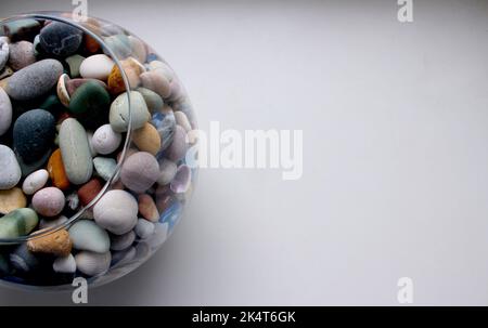 Concetto per una cartolina o sfondo con una superficie bianca e un vaso rotondo con pietre di mare e conchiglie sul lato Foto Stock