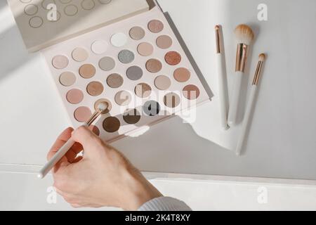 Mano prendendo cosmetici per il trucco, fiori di rose sulla tavola. Ombre decorative degli occhi con fondazione sul tavolo da toeletta nel salone di bellezza. Scelta dell'artista Foto Stock