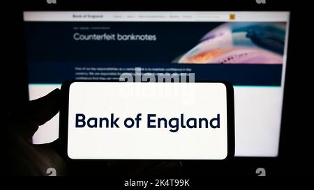 Persona che tiene il cellulare con il logo della Banca britannica emittente di Inghilterra sullo schermo di fronte alla pagina web. Messa a fuoco sul display del telefono. Foto Stock