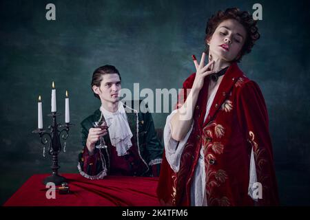 Ritratto di spaventosa famiglia di nobili vampiri, uomo e donna in costumi medievali che bevono sangue su sfondo verde scuro Foto Stock