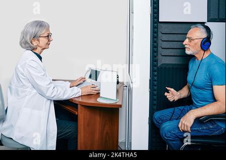 Esame audiologico. Audiologo che fa check-up uditivo a un uomo anziano in cabina audiometrica insonorizzata. Trattamento perdita dell'udito, vista laterale Foto Stock