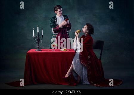 Ritratto di uomo e donna a immagine di vampiri su sfondo verde scuro. Uomo che prende selfie di donna che mangia hamburger Foto Stock