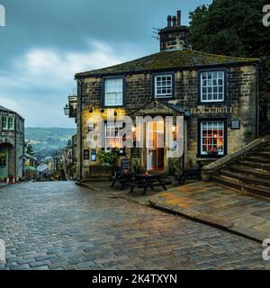 Haworth Main Street (collina ripida, vecchi edifici, luce notturna blu, storico villaggio delle sorelle Bronte, pub di grado 2) - West Yorkshire, Inghilterra, Regno Unito. Foto Stock
