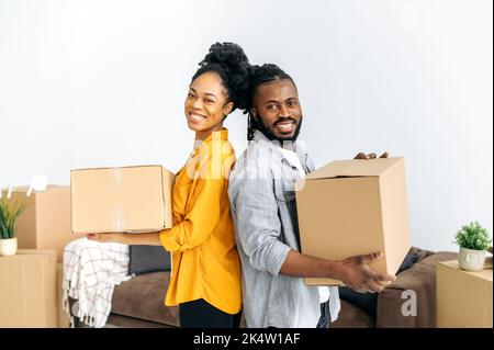 Felice moderno sposato afro americano coppia, tenendo scatole con le cose per la casa, in piedi nel loro nuovo alloggiamento, guardando la macchina fotografica, sorridendo, gioendo nell'acquisto del loro proprio appartamento o casa Foto Stock