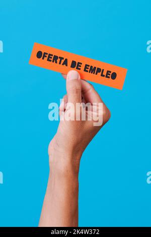 primo piano di un uomo che tiene un cartello di carta arancione con il testo di offerta di lavoro scritto in spagnolo, su uno sfondo blu Foto Stock