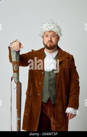 Studio shot di giovane uomo a immagine di medievale in vestito da caccia marrone vintage e parrucca bianca con vecchio fucile da caccia isolato su sfondo grigio. Foto Stock