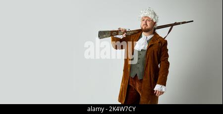 Cacciatore. Studio shot di giovane uomo a immagine di medievale in vestito da caccia marrone vintage e parrucca bianca con vecchio fucile da caccia isolato su grigio Foto Stock