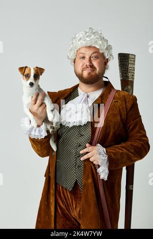 Cacciatore retrò con cane piccolo. Giovane uomo a immagine di persona medievale in vestito da caccia marrone vintage e parrucca bianca con vecchio fucile da caccia isolato Foto Stock