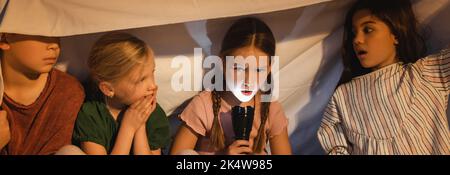 Ragazza che tiene torcia vicino amici spaventato sotto coperta a casa, banner, immagine stock Foto Stock