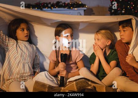 Ragazza che tiene la torcia vicino agli amici interracial spaventati sotto coperta e decorazione di natale a casa, immagine di riserva Foto Stock