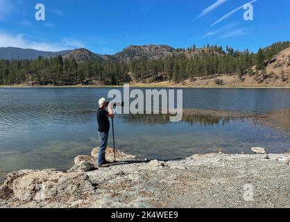 Fotografo in piedi sulla spiaggia utilizzando un teleobiettivo e un cavalletto, Okanagan Lake, Okanagan, British Columbia, Canada Foto Stock