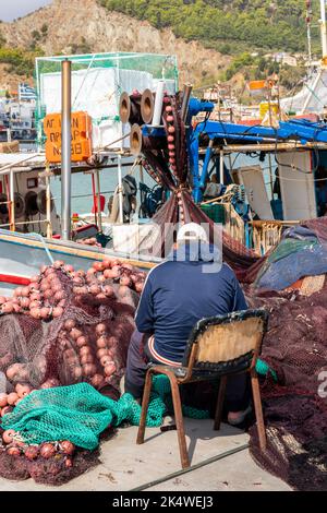 pescatore greco seduto in un porto vicino alla sua barca che ripara le sue attrezzature e le sue reti per la manutenzione Foto Stock