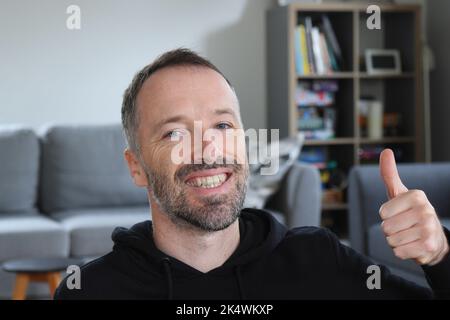 L'uomo sorride e mostra un gesto con i pollici. Uomo bearded con cappuccio nero casual. Uomo caucasico adulto di 30s o 40s anni. Feedback positivo approvazione gest Foto Stock