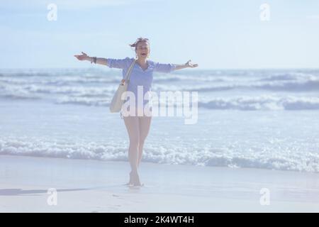 Ritratto a tutta lunghezza di felice donna elegante con sacco di paglia bianco che si rallegrano in spiaggia. Foto Stock