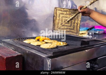 Primo piano della mano di una donna che cuoce il carbone dalla griglia per preparare le empanadas colombiane e gli arepas con un ventilatore Foto Stock