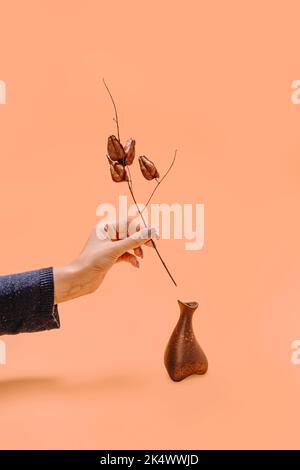 La mano di una donna tiene un rametto di fiori secchi di forma insolita. Forme insolite di marrone cioccolato su sfondo biondo autunnale. Semplice natura moderna ba Foto Stock