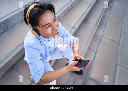 felice scatto di un uomo asiatico che ascolta musica con le cuffie mentre si siede sulle scale fuori e ridendo Foto Stock