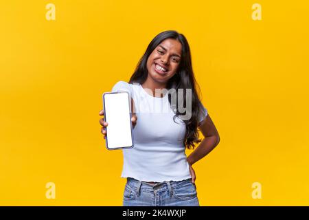 Donna indiana felice isolato guardando la macchina fotografica mentre mostra il telefono su uno sfondo giallo. Foto Stock