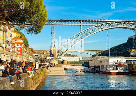 PORTO, PORTOGALLO - 7 NOVEMBRE 2021: Persone sedute al sole sull'argine di Ribera, ponte di Dom Luis sullo sfondo, sole serale, Porto, Portogallo Foto Stock