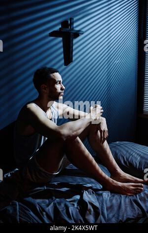 Un giovane depresso seduto sul letto mentre rimane sveglio tutta la notte fino alla mattina e guardando la finestra con le persiane veneziane Foto Stock