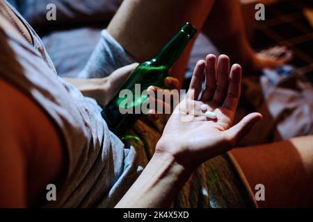Sopra l'angolo di mano di giovane depresso uomo che tiene le pillole di sonno o antidepressivi e bottiglia di alcol mentre si siede sul letto Foto Stock