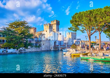 Il bellissimo porto di Sirmione con piccole imbarcazioni turistiche e da pesca contro le case colorate e Castello Scaligero, Lombardia, Italia Foto Stock