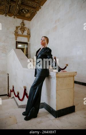 Donna piuttosto sottile che indossa un abito in pelle scura in piedi in un maestoso, storico interno, appoggiato su una parete di marmo Foto Stock