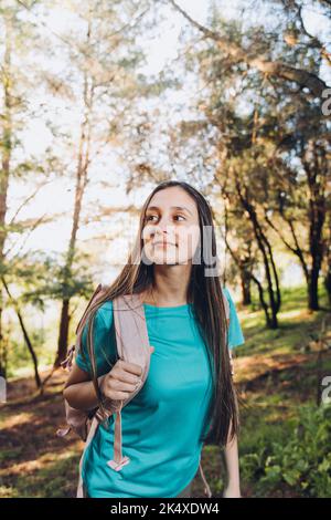 Fiducia in se stessi. Giovane donna con t-shirt turchese, zaino con sorriso sereno nella foresta. Verticale Foto Stock