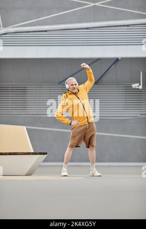 Ritratto verticale a tutta lunghezza di un uomo sportivo maturo che si estende all'aperto con cuffie in città Foto Stock