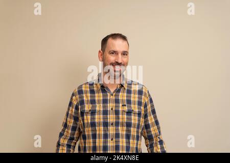 Ritratto dell'uomo ispanico bearded vestito in una camicia plaid che guarda la macchina fotografica mentre sorride, isolato su sfondo beige. Foto Stock