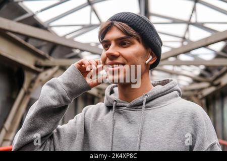 Giovane uomo hipster con auricolari wireless bianchi che ascoltano musica su sfondo urbano. Foto Stock