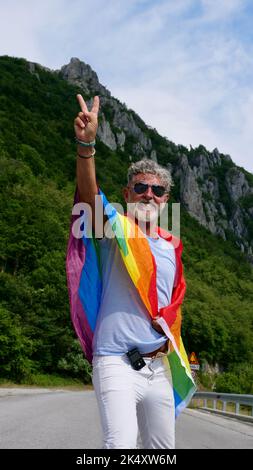 Ritratto di un anziano anziano dai capelli grigi bisessualità uomo caucasico con barba e occhiali da sole con bandiera di pace arcobaleno LGBTQIA in montagna. Celebra il mese dell'orgoglio, il giorno della bandiera dell'arcobaleno, la parata gay Foto Stock