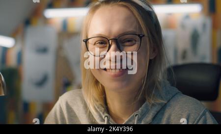 Dettagli di una giovane donna bionda con un nero occhiali incorniciati guarda la macchina fotografica e sorride. Donna si siede su una sedia in una configurazione basata domestica dell'ufficio. Foto Stock