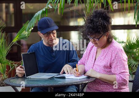 Uomo e donna maturi in occhiali che parlano tra loro, seduti a un tavolo, scrivere in blocco note vicino al computer portatile sotto la palma Foto Stock