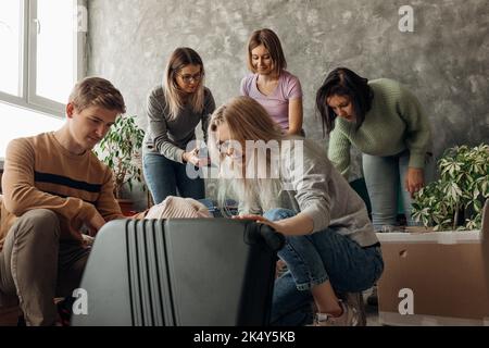 Amici sorridenti e affollati che si accollano i bagagli e cercano di mettere le cose in valigia, valigia. Trasferimento e trasferimento di appartamento Foto Stock
