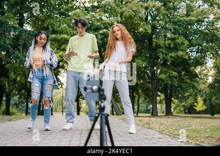 Gruppo di amici trendy che girano un vlog con uno smartphone montato su un cavalletto. Divertitevi a ballare nel parco cittadino. Ascoltare musica, senza stress. Foto Stock