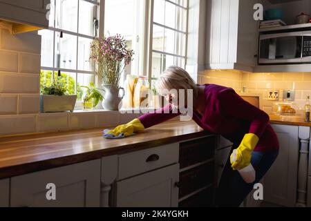 Donna causiana felice che indossa guanti di gomma, pulizia del banco in cucina Foto Stock