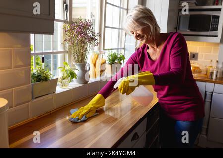 Donna causiana felice che indossa guanti di gomma, pulizia del banco in cucina Foto Stock