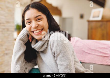 Ritratto di felice donna biraciale seduta sul divano in soggiorno, guardando la macchina fotografica e sorridendo Foto Stock